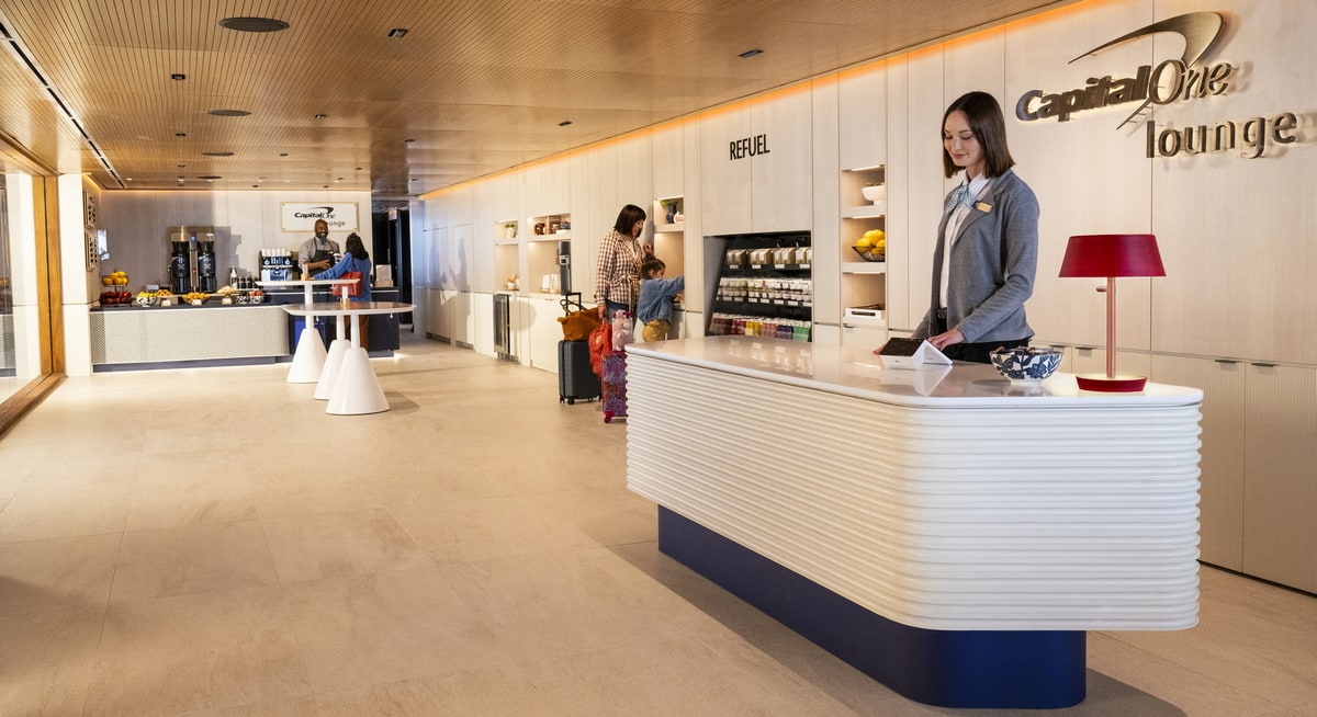Rendering of the Capital One lounge with a person standing at the desk and customers in the background.