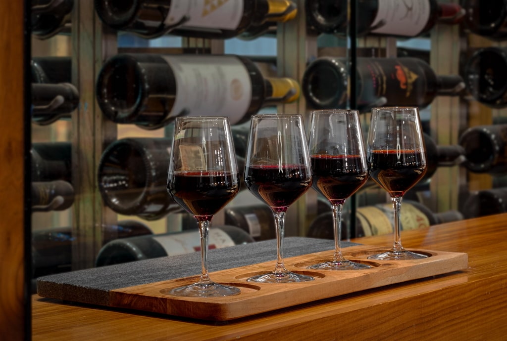 four filled wine glasses with bottles in the back on a table.