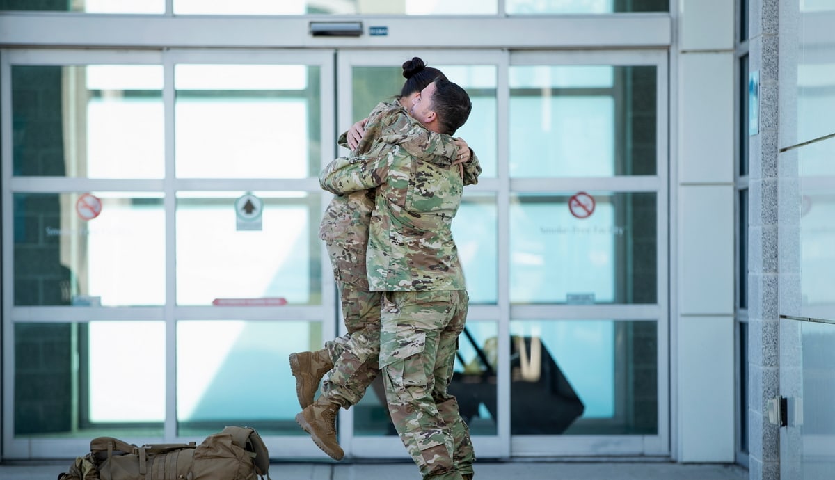 Two people in camouflage are hugging in celebration in front of the airport entrance doors.