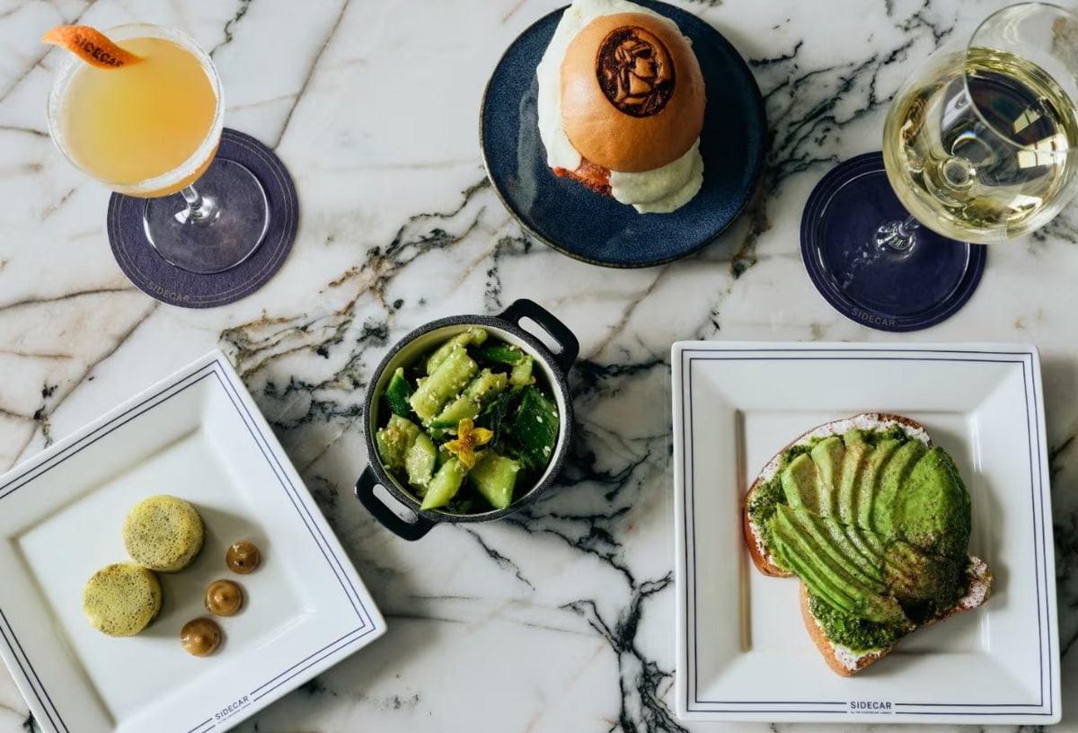 Sample menu items, including avacodo toast, crushed cucumber salad, wine and more on a marble table.