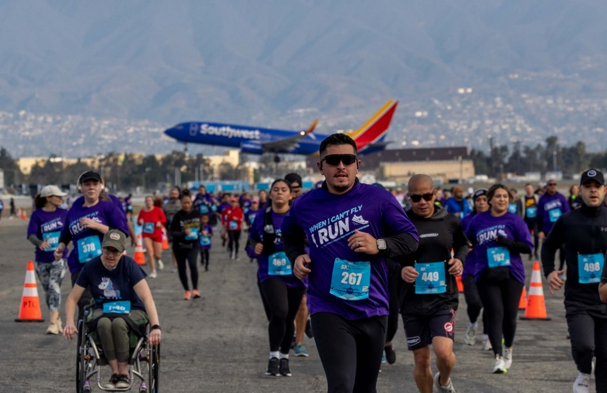 people competing on a runway run and someone in a wheelchair on the with a plane taking off in the background.